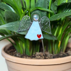 May include: A stained glass angel plant stake with a light blue body, textured wings, and a red heart. The angel has a silver halo and is set in a terracotta pot with green foliage.