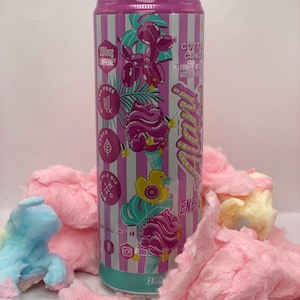 May include: A cylindrical candle designed to resemble an Alani Nu energy drink can with a pink and white striped label. The label features graphics of cotton candy, a yellow rubber duck, and text including 200mg caffeine and cotton candy.