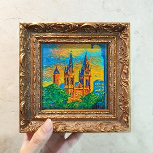 May include: A framed oil painting of a cathedral with two tall spires, set against a blue and yellow sky. The artwork is in a gold ornate frame. Green trees are at the base of the cathedral.