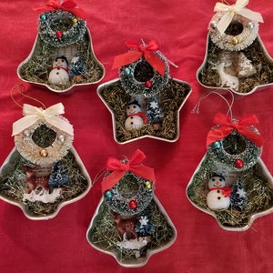 May include: Six Christmas ornaments made from cookie cutters. The ornaments are shaped like bells and stars. Each ornament is filled with white and green tinsel, a small Christmas tree, a snowman, a reindeer, and a red or white bow.