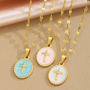 May include: Three gold-colored necklaces with oval pendants. Each pendant features a cross design in the center, surrounded by a colored border. The colors are light blue, pink, and white. The necklaces have delicate chains.