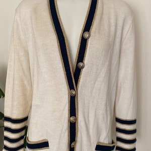 May include: A white cardigan with navy blue and gold trim. The cardigan has a V-neck and button closure. The sleeves have navy blue and gold stripes.