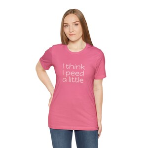 Puede incluir: Una camiseta rosa con el texto "I think I peed a little" en blanco. La camiseta tiene cuello redondo y mangas cortas. La modelo lleva vaqueros azules.