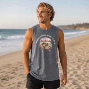 A man on a beach wears a tank top with a graphic of a frog working on a laptop in a van.