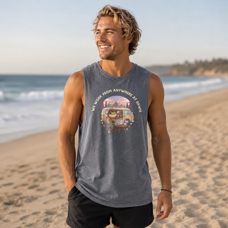 A man on a beach wears a tank top with a graphic of a frog working on a laptop in a van.