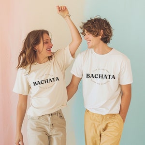 May include: Two people wearing white t-shirts with the text "Life is better when I'm dancing Bachata" printed on them. The people are smiling and dancing.
