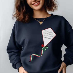 Può includere: Felpa blu navy con un design a forma di aquilone bianco con un nastro rosso, bianco e verde. L'aquilone contiene una citazione: "Se devo morire, che porti speranza, che sia una storia" di Refaat Alareer.