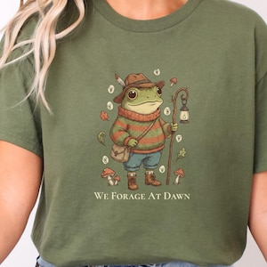 Op de afbeelding: Een olijfgroen t-shirt met een afbeelding van een kikker die een hoed, trui en laarzen draagt, met een tas en een lantaarn aan een staf. De illustratie bevat kleine paddenstoelen en bladeren, met de tekst "WE FORAGE AT DAWN" eronder gedrukt.