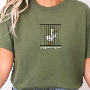 May include: An olive green t-shirt featuring a graphic of a white goose with an orange beak and feet standing inside a black cage. The goose holds a small spoon in its wing. Below the cage, the text reads, "guilty of monitoring my spoons."