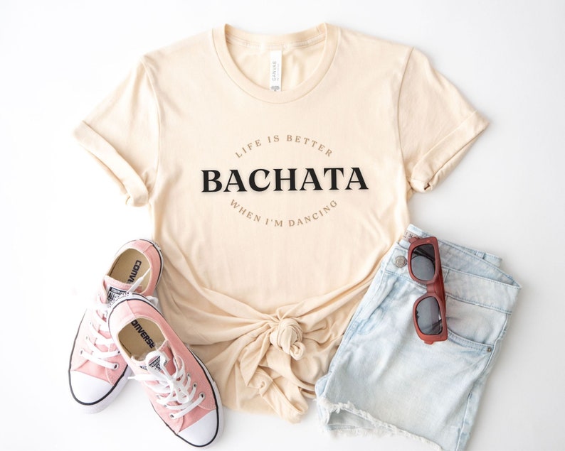 May include: A beige t-shirt with the text "Life is better when I'm dancing Bachata" printed on the front. The shirt is paired with pink sneakers and denim shorts.