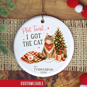 May include: A round white ceramic Christmas ornament featuring a brown tabby cat wearing a red bandana, a decorated Christmas tree, wrapped gifts, and a mug of cocoa. The text reads Plot Twist... I GOT THE CAT, Francesca, and 2026.