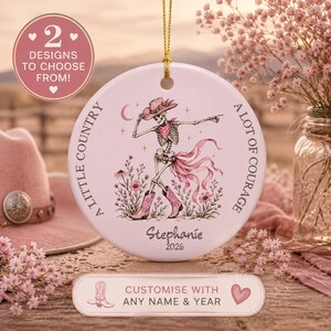 May include: A round white ceramic ornament featuring a skeleton wearing a pink cowboy hat, bandana, and boots, surrounded by flowers. The text reads A LITTLE COUNTRY A LOT OF COURAGE, Stephanie, and 2026. It hangs from a gold string.