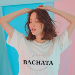 May include: A woman wearing a white t-shirt with the text "Life is better when I'm dancing Bachata" printed on it. She is standing in front of a colorful background.
