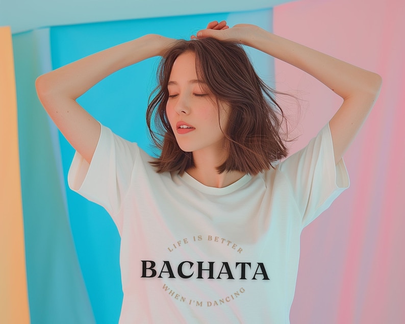 May include: A woman wearing a white t-shirt with the text "Life is better when I'm dancing Bachata" printed on it. She is standing in front of a colorful background.