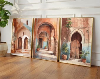 Può includere: Set di tre stampe artistiche incorniciate raffiguranti scene ad acquerello di architettura mediorientale. L'opera presenta porte ad arco, passaggi ed edifici in toni caldi e terrosi con cornici dorate. Le piante in vaso aggiungono un tocco di verde.