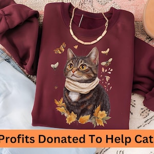 Può includere: Una felpa color borgogna con una grafica di un gatto che indossa una sciarpa e circondato da foglie autunnali e farfalle. Il testo "Profits Donated To Help Cats!" è nella parte inferiore dell'immagine.