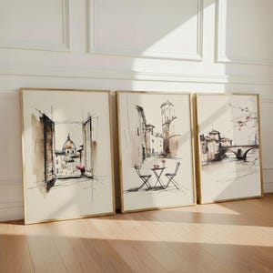 以下が含まれることがあります： イタリアの街並みをインクと水彩で描いたスケッチの額入りアートプリント3枚セット。左は開いた窓から見えるドーム型の建物、中央は椅子が2脚あるストリートカフェ、右は石造りの橋が描かれています。