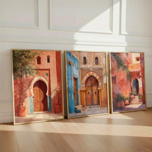 Puede incluir: Tres impresiones artísticas enmarcadas que representan escenas vibrantes de la arquitectura marroquí. La obra presenta puertas arqueadas, puertas de madera y paredes coloridas en tonos rojos, azules y naranjas. Las impresiones se exhiben sobre un suelo de madera.