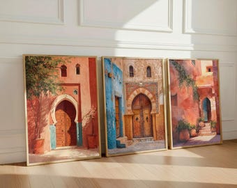 May include: Three framed art prints depicting vibrant scenes of Moroccan architecture. The artwork features arched doorways, wooden doors, and colorful walls in shades of red, blue, and orange. The prints are displayed on a wooden floor.