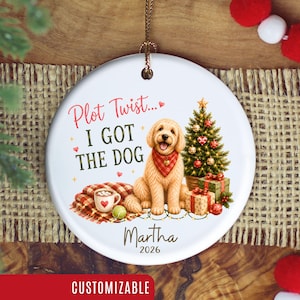 Puede incluir: Un adorno navideño redondo de cerámica blanca con un perro golden doodle con pañuelo de cuadros rojos, un árbol de Navidad decorado, regalos y una taza de cacao. El texto dice Plot Twist... I GOT THE DOG, Martha, 2026.