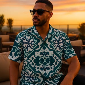 May include: A short-sleeved button-down shirt with a teal and white abstract pattern. The shirt is worn with black sunglasses. The background features a sunset and string lights, suggesting an outdoor setting.