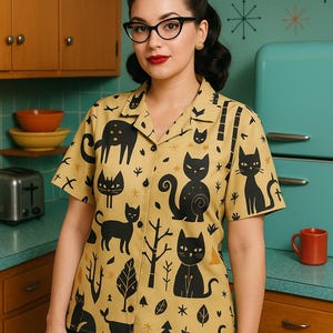 May include: A mustard yellow button-down shirt featuring a black cat design. The short-sleeved shirt has a classic collar. The person wearing the shirt is in a retro kitchen with a turquoise refrigerator.