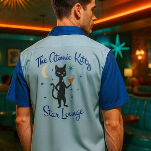 May include: A light blue and navy blue short-sleeved shirt with a retro design. The back of the shirt features the text "The Atomic Kitty Star Lounge" with a black cat illustration holding a cocktail.