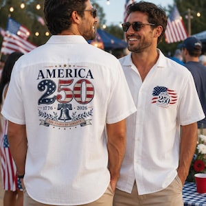 May include: A white short-sleeved button-down shirt featuring a graphic on the back that reads AMERICA 250, 1776-2026, and 250 YEARS OF LIBERTY with a Liberty Bell illustration. A smaller version of this graphic is printed on the front left chest.