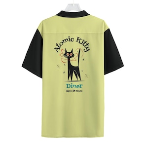 May include: A yellow and black short-sleeved bowling shirt with a retro design. The shirt features a black cat with red eyes in a circle of stars and the text "Atomic Kitty Diner Open 24 Hours".