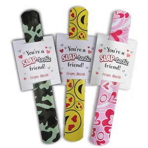 May include: Three slap bracelets with attached cards. The bracelets have different patterns: camouflage, yellow smiley faces, and pink hearts. Each card reads "You're a SLAP-tastic friend!" and "From: Alecia."