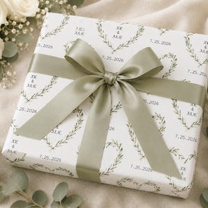 May include: A square gift box wrapped in white paper featuring a repeating pattern of green leaf wreaths, the names Joe and Julie, and the date 7.25.2026. The box is tied with a wide, smooth sage green satin ribbon tied in a large bow on top.