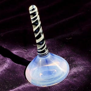 Puede incluir: Un tapón de botella de vidrio transparente con una base bulbosa y un tallo largo en espiral. El tallo presenta rayas negras y blancas alternas. La base tiene un efecto iridiscente azul y blanco.