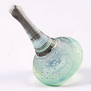 May include: A clear glass bottle stopper with a rounded base and a long, angled stem. The base is filled with small bubbles and has a light blue and green iridescent finish. The stem is a dark gray color.