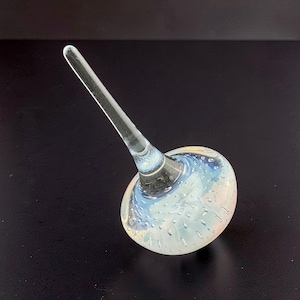 May include: A clear glass bottle stopper with a long, slender stem and a rounded base. The base features a swirl of blue and white colors with small air bubbles, creating a decorative effect. The stopper is designed to fit into a bottle.