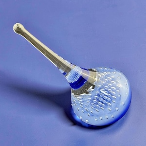 May include: A clear glass bottle stopper with a blue bulbous base and a long, clear stem. The base is decorated with small, bubble-like details and blue accents. The stopper is approximately 6 inches long.