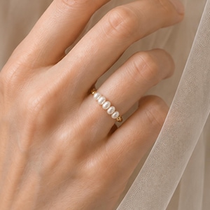 Op de afbeelding: Een gouden ring met een rij van vijf kleine, witte, ovaalvormige zoetwaterparels. De parels zijn horizontaal gerangschikt langs de dunne, gepolijste gouden band, wat zorgt voor een verfijnde en delicate uitstraling aan de vinger.