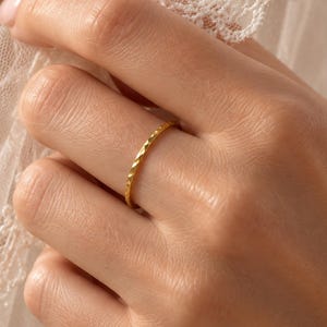 Puede incluir: Un anillo fino de oro amarillo con una superficie texturizada y facetada. La banda presenta un patrón geométrico repetitivo que capta la luz, creando un brillo sutil. El anillo se lleva en un dedo, parcialmente cubierto por encaje blanco.