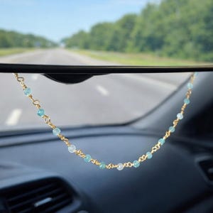 May include: A gold-colored chain with light blue beads hangs from a car's rearview mirror. The mirror reflects a sunny day on a highway with green trees on either side. The car's dashboard is visible.