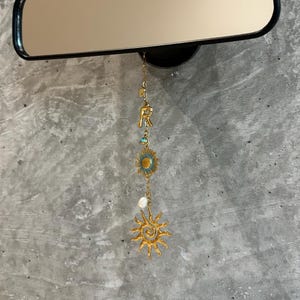 Può includere: Uno specchietto retrovisore con cornice nera con un ciondolo decorativo appeso sotto. Il ciondolo presenta elementi color oro tra cui la lettera "R", un girasole e un disegno a raggiera, accentuato con turchese e una perla.