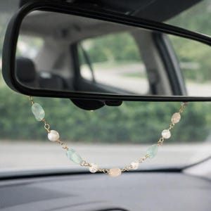May include: A car rearview mirror with a decorative beaded chain hanging from it. The chain features a mix of pale green, white, and peach-colored beads strung on a gold-colored chain. The car's interior is visible in the background.