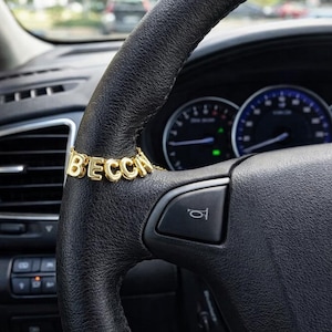 Puede incluir: Un collar dorado con el nombre "BECCA" cuelga sobre un volante de cuero negro. El collar está formado por letras doradas individuales unidas por una fina cadena. El salpicadero y el interior del coche son visibles en el fondo.
