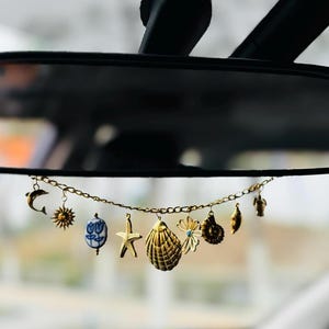 Könnte beinhalten: Eine goldene Kette mit verschiedenen Anhängern, darunter eine Muschel, ein Seestern, eine Blume, eine Schildkröte, ein Delfin und eine Sonne. Die Kette hängt am Rückspiegel eines Autos.