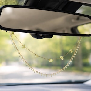 May include: A car's rearview mirror with a decorative hanging ornament. The ornament features two strands of gold-colored chain, adorned with small, light-green heart-shaped beads and tiny white beads. The background shows a blurred view of a road and trees.