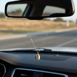 May include: A gold chain with a seashell charm hangs from a car's rearview mirror. The seashell is beige and oval-shaped. The car's dashboard is visible, with a blurred road and landscape in the background.