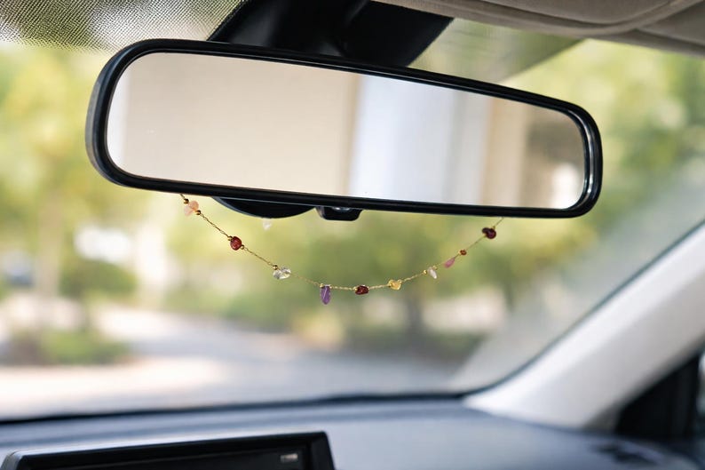 May include: A car's rearview mirror with a black frame, reflecting a blurred outdoor scene. A delicate gold chain hangs below the mirror, adorned with colorful gemstones. The gemstones include red, pink, purple, and clear crystals.