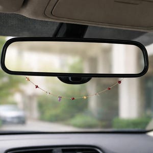 May include: A car's rearview mirror with a decorative beaded garland hanging below it. The garland features small red, purple, and yellow beads strung on a gold-colored chain. The background shows a blurred outdoor scene.