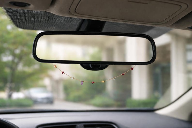 May include: A car's rearview mirror with a decorative beaded garland hanging below it. The garland features small red, purple, and yellow beads strung on a gold-colored chain. The background shows a blurred outdoor scene.