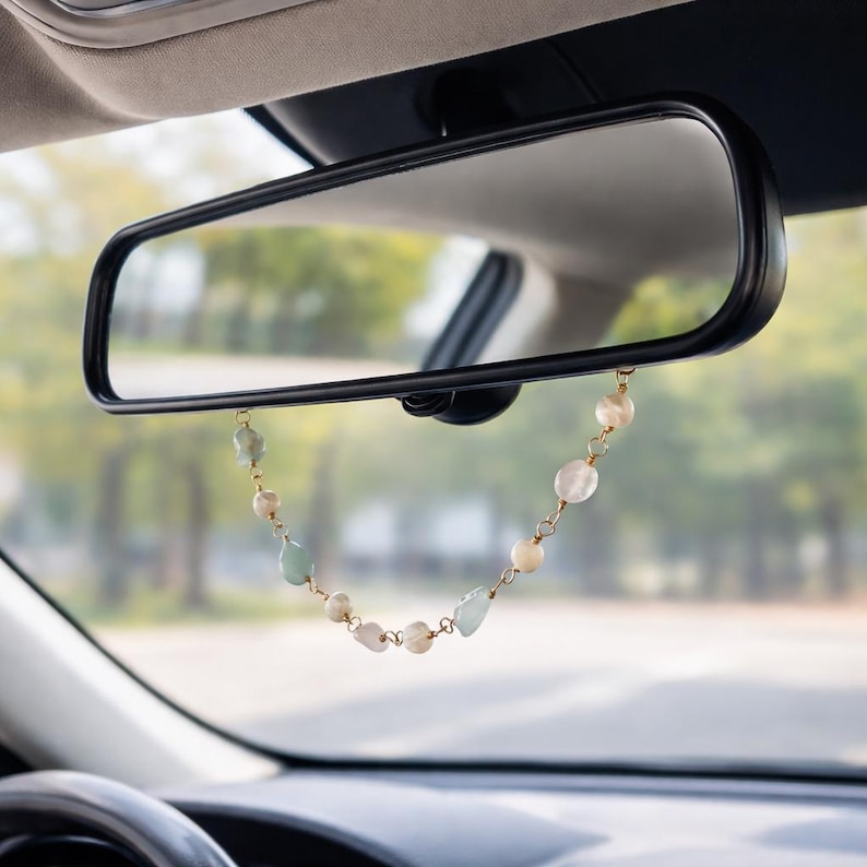 May include: A car rearview mirror with a decorative beaded charm hanging from it. The charm features a mix of light green, white, and gold-colored beads. The background shows a blurred view of a road and trees.