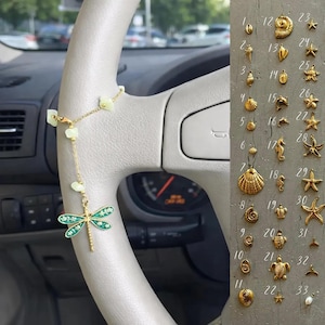 May include: A gold-coloured car charm with a dragonfly pendant and a chain of green beads is attached to a car steering wheel. A collection of gold-coloured seashell charms are displayed on a grey surface.