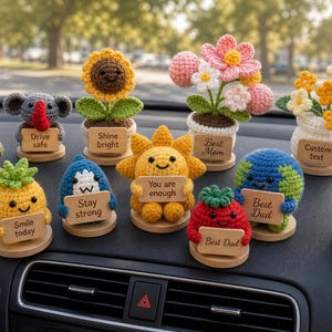 May include: A collection of crocheted car accessories, including a turtle, elephant, sunflower, pineapple, shark, sun, tomato, earth, flowers, and broccoli. Each figure holds a sign with positive messages like "You got this" and "Smile today."